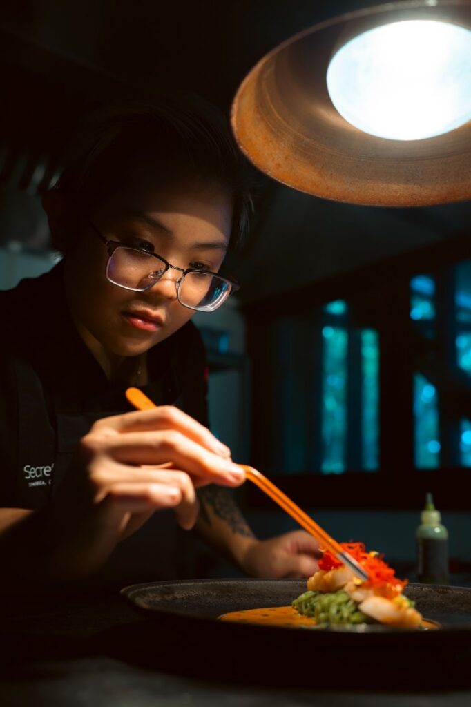 Chef Allison Lo under a kitchen light putting the final ingredients on a colorful dish at Zing Zing at Secret Bay Dominica.
