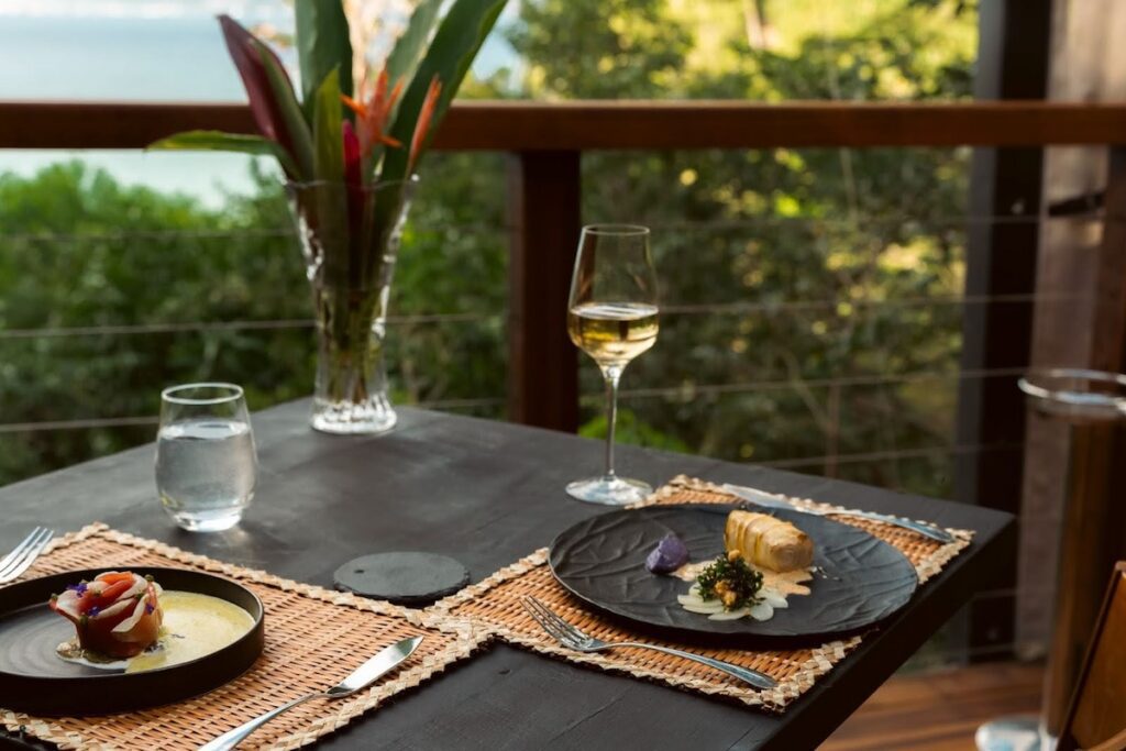 A table set for 2 with entrees and wine facing toward Tibay Beach from the patio at Zing Zing, Secret Bay Dominica