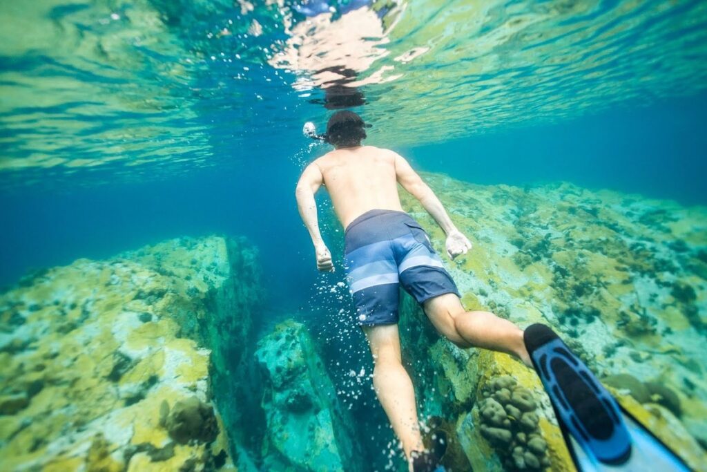 Man snorkeling in clear blue water above rocky coral formations, swimming with fins toward an underwater reef.