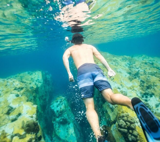 Man snorkeling in clear blue water above rocky coral formations, swimming with fins toward an underwater reef.